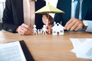 Insurance agents holding a small umbrella over paper cutouts of a family and home.