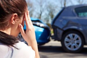 A woman holds her hand to her temple and wonders, “Does auto insurance cover rental cars?” as she looks at the damage from a rear-end collision.